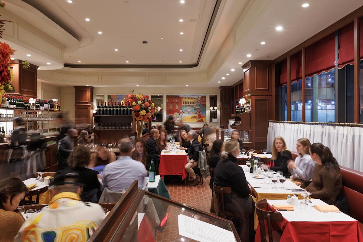 A lively restaurant scene with people dining at tables, chatting, and enjoying drinks. The interior features warm lighting, wooden details, large windows, and floral centerpieces adding color to the elegant setting.