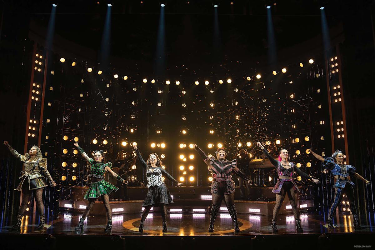 Six performers in colorful, historical costumes sing and pose energetically on a lit stage, with spotlights and glitter in the background, creating a lively, theatrical scene.