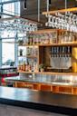 A modern bar with shelves of assorted liquor bottles, beer taps, hanging glasses, and potted plants, set against a wooden and metal backdrop with natural light coming through large windows.