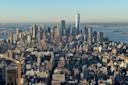 Aerial view of a cityscape featuring numerous skyscrapers and buildings under a clear blue sky. Prominent structures include a tall tower in the center. The distant horizon shows a body of water with a bridge and land beyond. Construction cranes visible.