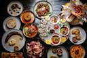 A variety of gourmet dishes on a dark table, including oysters with sauces and lemon, seafood, roasted meats, salads, grilled octopus, flatbread, and assorted appetizers arranged artfully on plates.