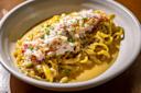 A close-up of a bowl of fresh pasta topped with shredded crab meat, garnished with chopped chives and golden breadcrumbs, served in a creamy yellow sauce.