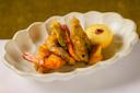 A scalloped white dish holds several pieces of crispy fried shrimp and vegetables, garnished with a lemon half filled with dipping sauce. The dish is placed on a white surface against a green background.