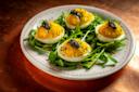 Four deviled eggs topped with caviar and herbs are arranged on a bed of arugula, served on a white plate with a pink decorative rim, sitting on a copper-colored surface.
