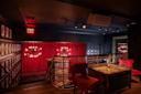 A dimly lit lounge with red curtains, red chairs, a wooden table, chandeliers, and framed photos on the wall. An EXIT sign glows above a stairway with a gold railing. The ambiance is cozy and intimate.