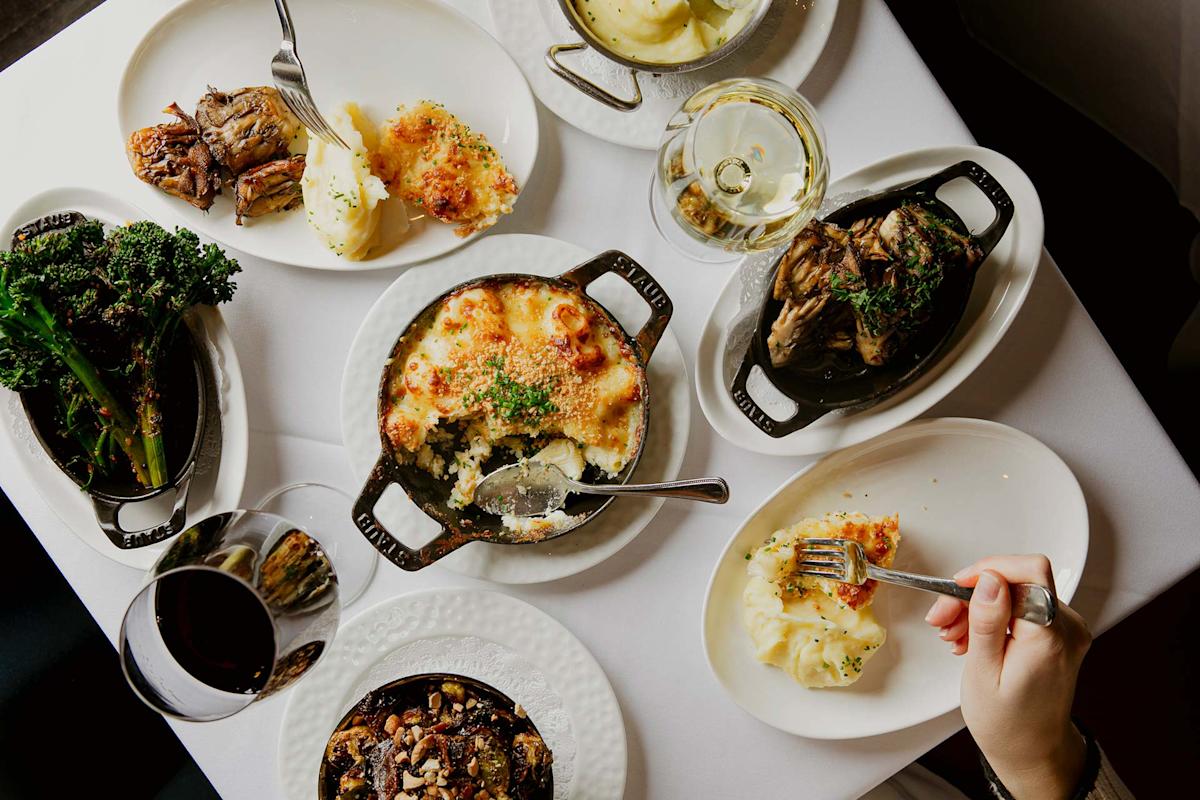 A table set with various dishes, including mashed potatoes, baked pasta, roasted vegetables, and glasses of red and white wine. A hand is holding a fork with mashed potatoes on one plate.