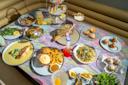 A table with a variety of colorful dishes, including seafood, fries, salad, pastries, drinks, and desserts, set on a pastel marble-patterned tablecloth in a modern dining setting.