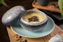 A ceramic bowl with a speckled lid, partially open, reveals a creamy yellow soup topped with a small mound of dark, chopped garnish. The bowl sits on a matching plate, with Asian calligraphy paper underneath.