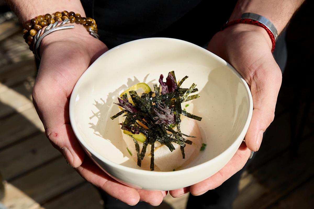 A person holds a white bowl containing a dish garnished with seaweed strips, leafy greens, and lemon slices. The person is wearing a beaded bracelet and a red string bracelet on their wrist. The background is softly blurred.