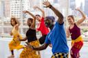 Five people of diverse backgrounds smile and dance energetically in a bright studio with city buildings visible through large windows. They wear colorful clothes and appear to be following a lively group choreography.