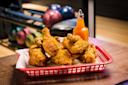A red basket lined with paper holds several pieces of crispy fried chicken on a wooden table. Behind it, there is a bottle of orange sauce and blurry bowling balls and lanes in the background.