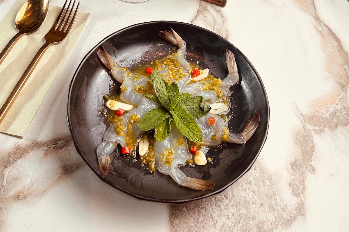 A black plate of raw shrimp garnished with mint leaves, sliced garlic, red chili, and a light sauce, placed on a marble table next to gold utensils and a beige napkin.