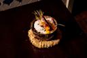 A dessert served in a coconut shell bowl, topped with whipped cream, spun sugar, edible flower petals, and a cinnamon stick, placed on a woven mat atop a dark wooden surface.