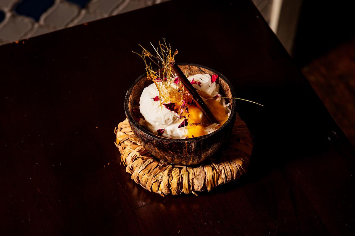 A dessert served in a coconut shell bowl, topped with whipped cream, spun sugar, edible flower petals, and a cinnamon stick, placed on a woven mat atop a dark wooden surface.