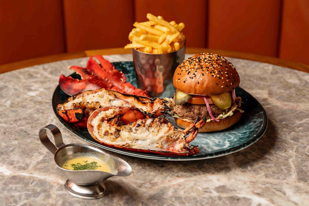 A gourmet platter featuring a sesame seed burger with pickles and onions, a side of fries in a metal cup, grilled lobster halves, and a small gravy boat of sauce, all served on a marble table.