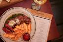 A plate with sliced steak topped with herb butter, thick-cut fries, and a side of green sauce. A small dish of ketchup is next to the fries. A glass of white wine and a menu are partially visible on the table.