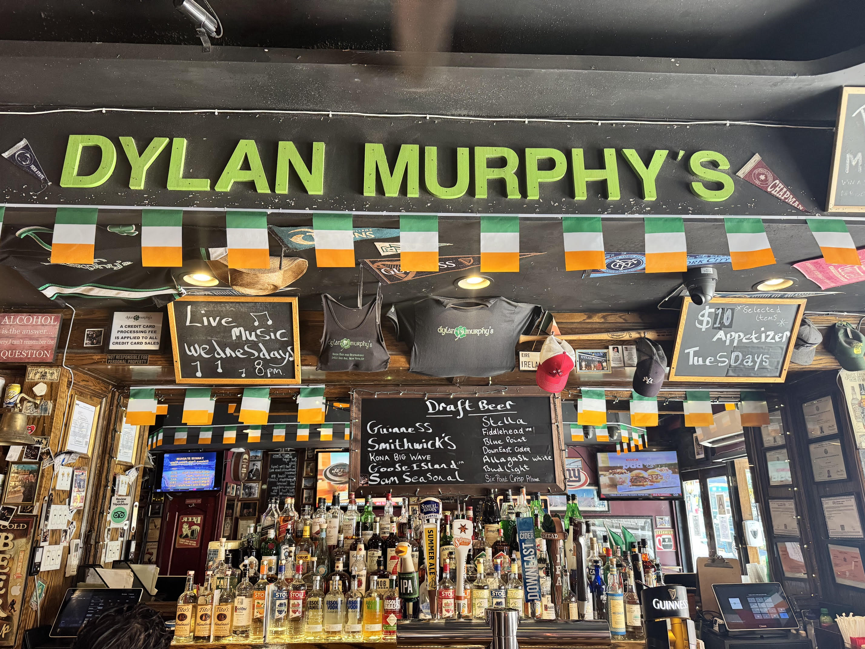 Bar interior at Dylan Murphy’s with Irish flags, liquor bottles, chalkboard menus listing draft beers and appetizer specials, T-shirts, and hats displayed above the bar. Signs advertise live music and weekly deals.