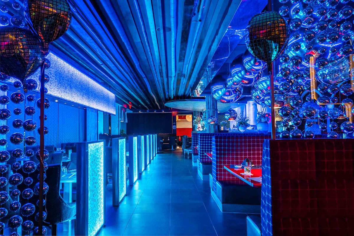 A vibrant, modern restaurant interior with blue neon lighting, reflective surfaces, disco balls, and red-accented booth seating, creating a futuristic and lively atmosphere.