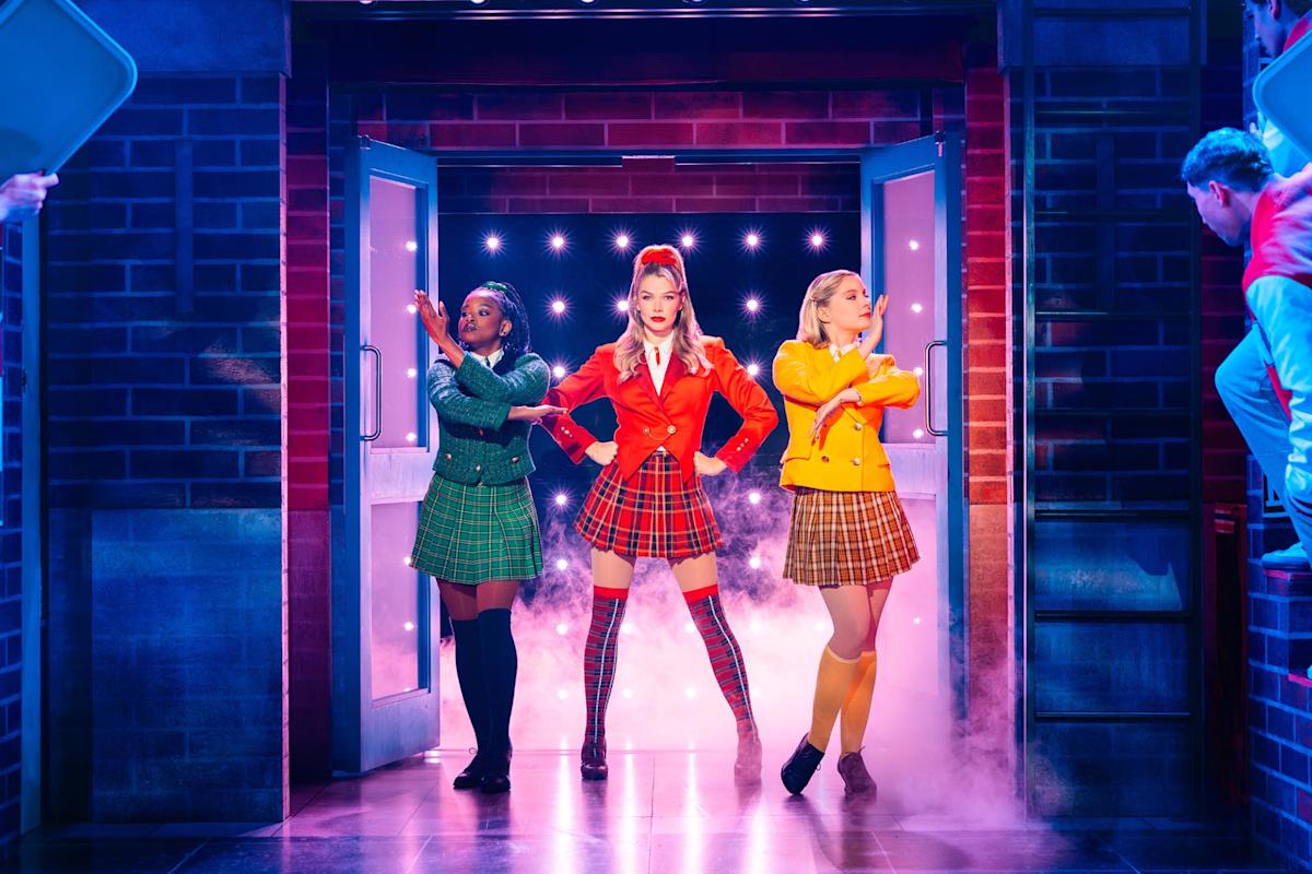 Three women in plaid skirts and colorful blazers—green, red, and yellow—pose confidently on stage with bright lights and fog behind them, as audience members watch from the sides.