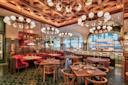 A stylish restaurant interior with wooden tables and red cushioned seating. Round chandeliers hang from a geometric wooden ceiling. Large windows provide natural light, and the bar area is visible in the background.