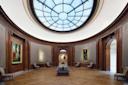 A circular gallery with wood-paneled walls, ornate doorways, classic paintings, upholstered chairs, and a large domed skylight illuminating the parquet floor and central benches.