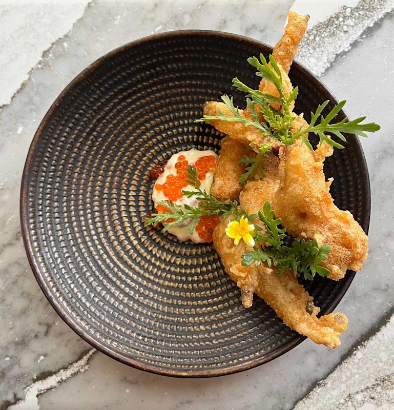A patterned bowl with fried frog legs garnished with greens and a yellow flower, served on a dollop of creamy sauce topped with orange fish roe, on a marble surface.