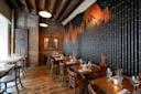 Cozy restaurant interior with wooden tables and chairs, exposed brick walls, wooden ceiling beams, and a unique black and orange textured wall design. The space is softly lit with warm hanging lights and candles on the tables.