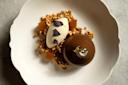 A white plate with a glossy chocolate dome topped with gold leaf, a quenelle of cream garnished with dried flower petals, crunchy granola pieces, and small golden jelly cubes, arranged artfully.