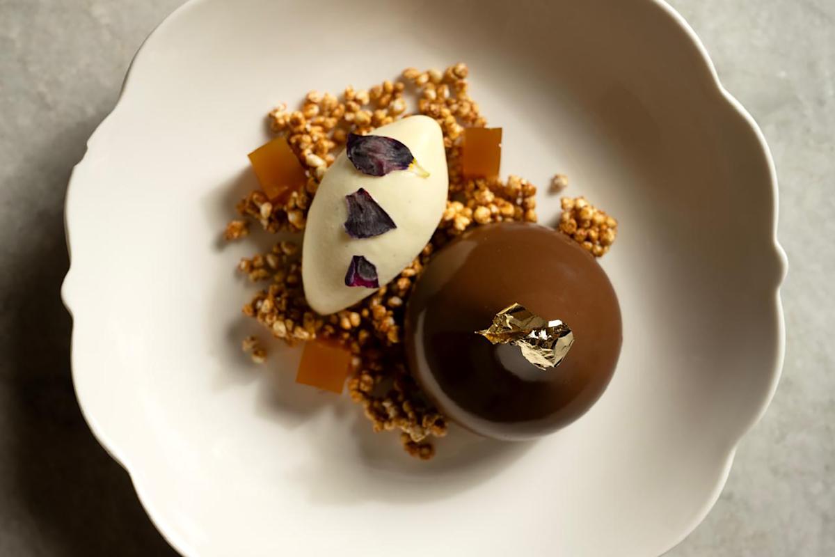 A white plate with a glossy chocolate dome topped with gold leaf, a quenelle of cream garnished with dried flower petals, crunchy granola pieces, and small golden jelly cubes, arranged artfully.