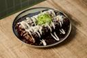 Two enchiladas covered in rich mole sauce, topped with crema, crumbled cheese, chopped onions, and shredded lettuce, served on a rustic green-rimmed plate on a wooden table.