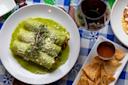 Three green enchiladas topped with microgreens and cheese on a white plate, served with green sauce. Next to it are tortilla chips with salsa and a glass of red wine on a blue checkered tablecloth.