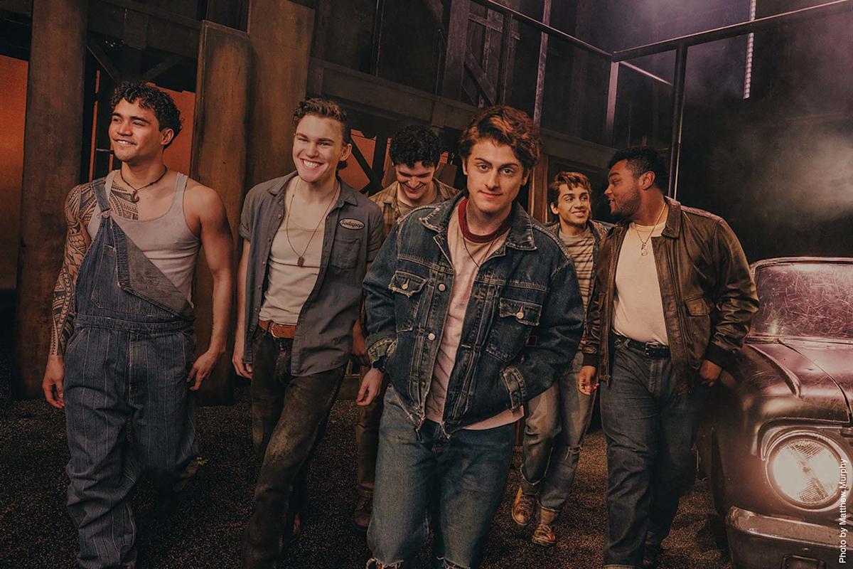 Six young men in casual 1950s-style clothing walk together, smiling, next to a vintage car in a dimly lit, industrial setting.