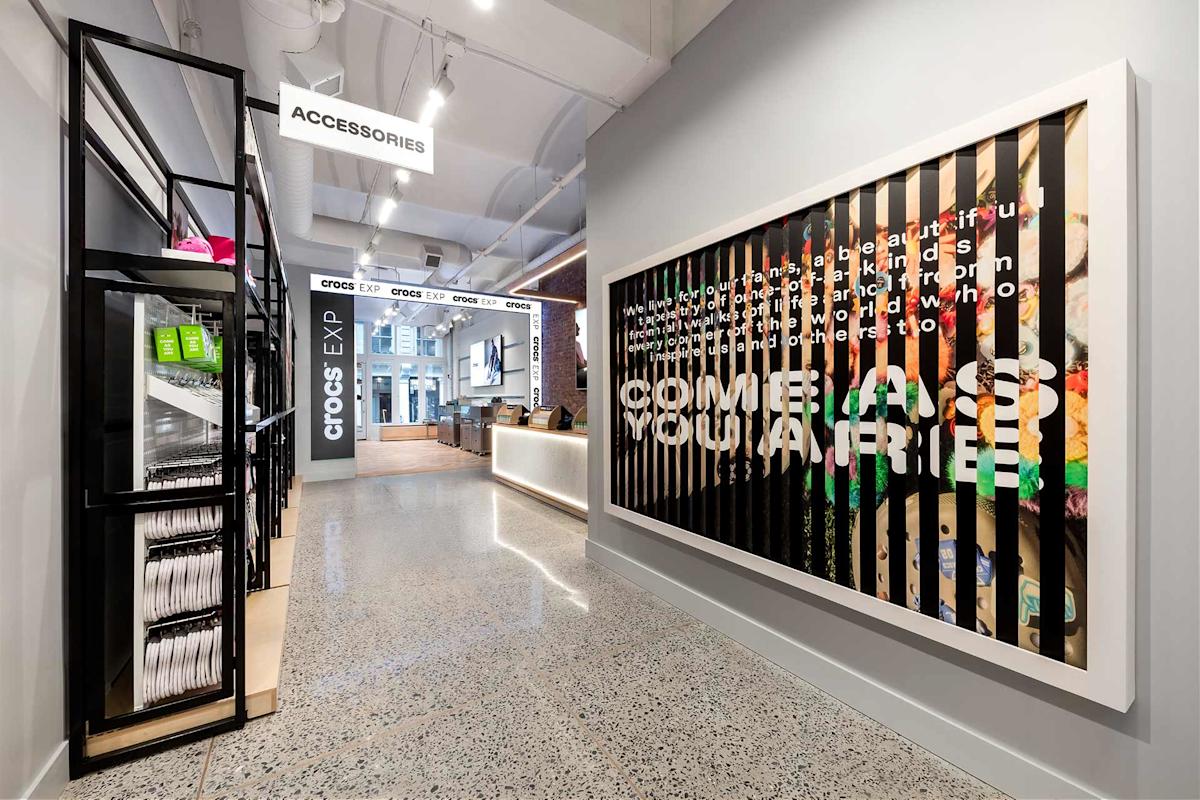 Modern retail store with high ceilings, bright lighting, a large “Accessories” sign, organized shelves, and a colorful Crocs display wall featuring a bold, partially obscured slogan. Reception counter visible in the background.