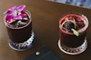 Two cocktails in glass tumblers on a table. The left drink is garnished with a purple orchid, and the right drink has a blackberry and dried citrus slice with a salted rim. Both are dark red in color.