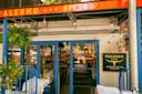 Exterior of Palermo Argentinian Bistro with a glass door, blue window frames, hanging lights, a green plant, and a yellow sign displaying the restaurant name on a white brick wall. Tables are set on the patio.