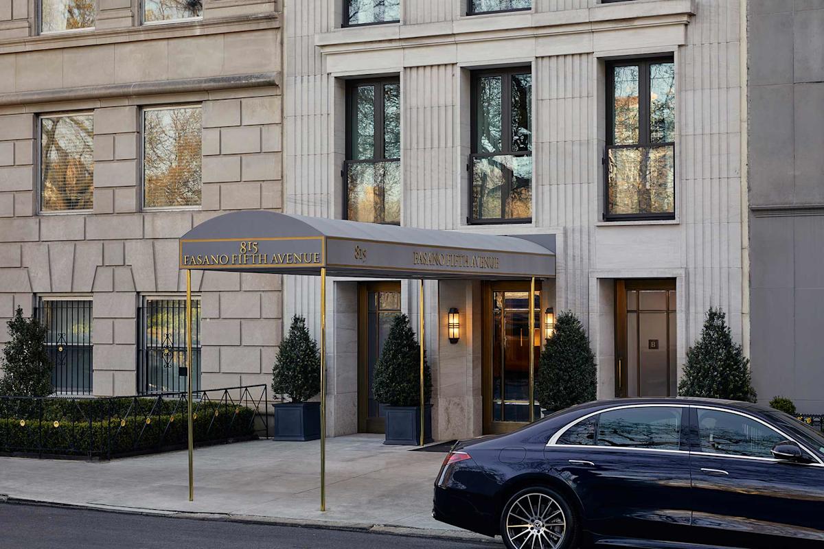 The entrance of a building with a gray canopy labeled "85 East 80th / 85 East 80th Avenue," flanked by potted plants. A black luxury car is parked on the street in front.