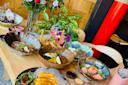 A table set with assorted Japanese dishes, including sashimi, noodles, grilled fish, and various sides, surrounded by flowers and decorative plants in a warmly lit restaurant setting.