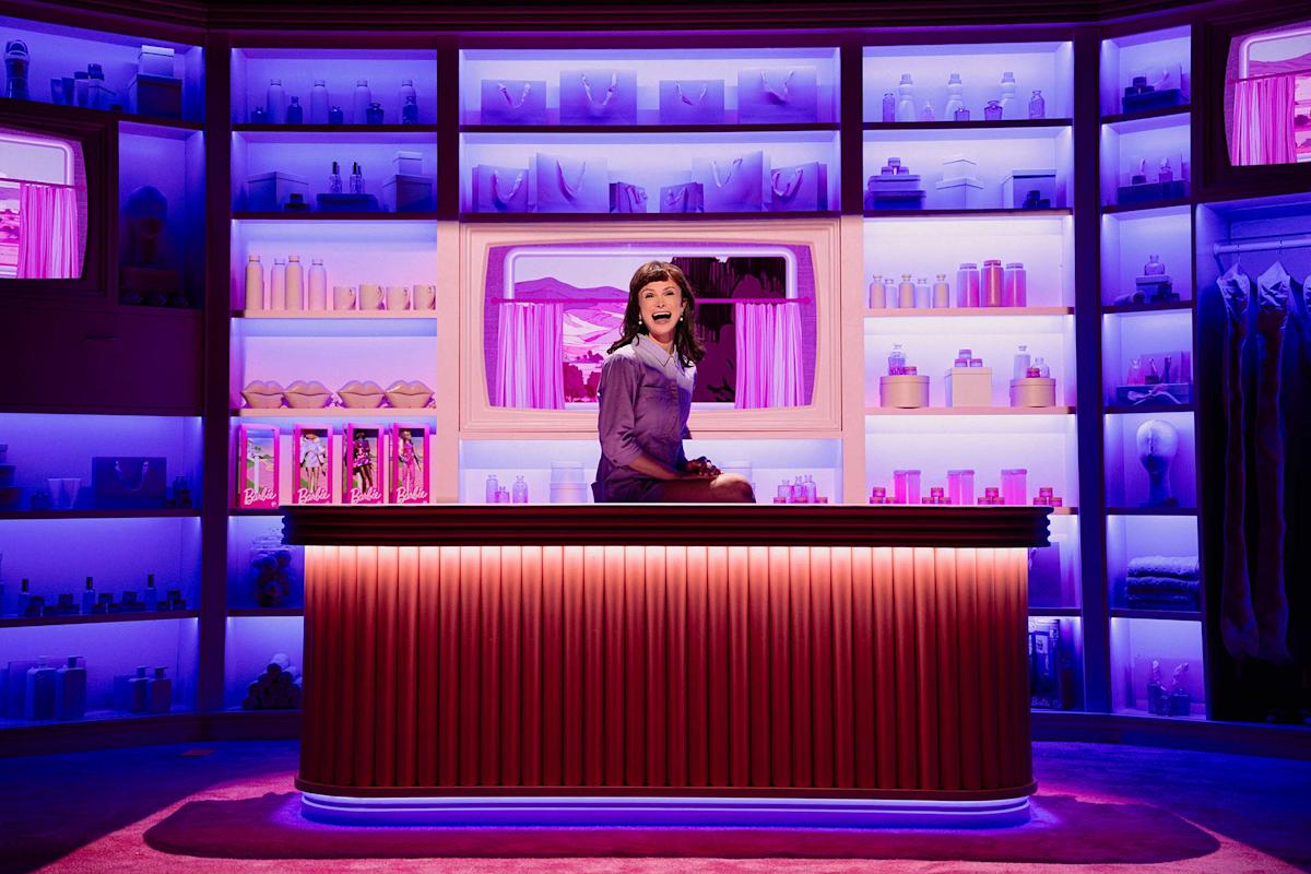 A woman in a purple outfit sits on a counter in a brightly lit room filled with assorted bottles and products on shelves. The lighting is mostly purple and pink, creating a vibrant, playful atmosphere.