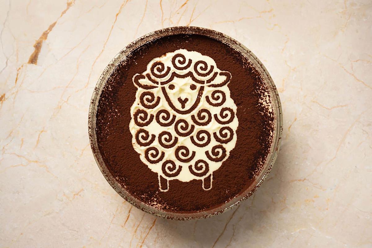 A bowl of dessert dusted with cocoa powder, featuring a cute sheep design made with cream and swirling patterns on top, set on a light marble surface.