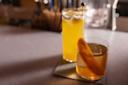 Two cocktails on a bar counter: one in a tall glass with a frothy top, and the other in a short glass garnished with a long orange peel. They sit on square coasters. The background is softly blurred.