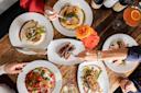 Overhead view of four people dining, sharing plates of gourmet food with vegetables, wine, and cocktails on a rustic wooden table, decorated with orange and yellow roses.