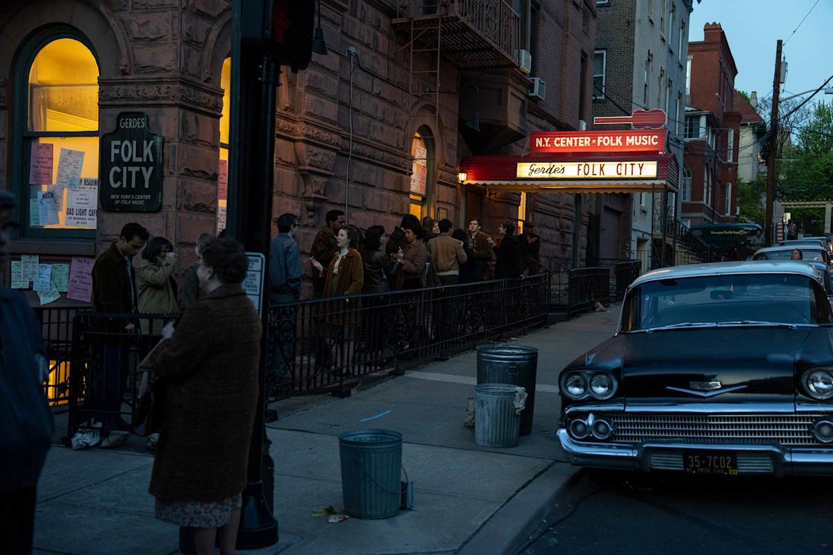 A crowd gathers outside "Folk City," a music venue with a lit marquee in the evening. People line up along the sidewalk, and an old-fashioned car is parked on the street. 