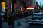 A crowd gathers outside "Folk City," a music venue with a lit marquee in the evening. People line up along the sidewalk, and an old-fashioned car is parked on the street. 