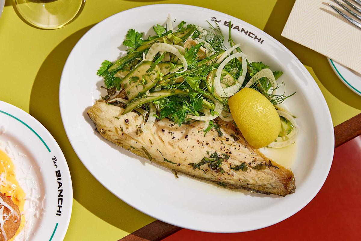 A white plate labeled "Bar Bianchi" holds a cooked fish fillet topped with fresh herbs, fennel, and onion, garnished with a lemon wedge. The plate is set on a yellow table next to a napkin and another dish.