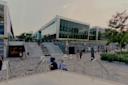 Modern university building with large glass windows, outdoor steps, people walking and sitting, and greenery around the entrance on a paved plaza. The sky is cloudy and the scene is lively with activity.