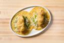 Two crispy battered fish tacos topped with shredded cabbage and cheese, served on corn tortillas on a white oval plate, placed on a light wooden table.