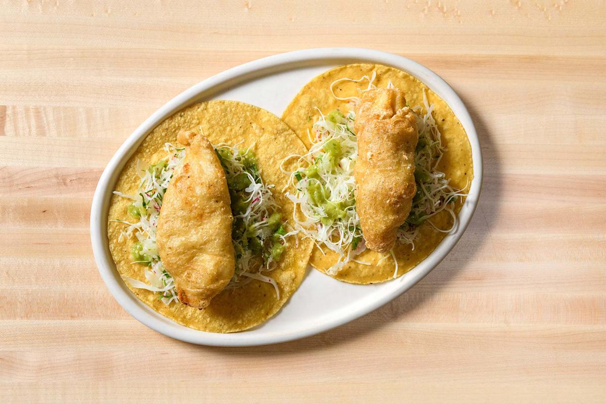 Two crispy battered fish tacos topped with shredded cabbage and cheese, served on corn tortillas on a white oval plate, placed on a light wooden table.