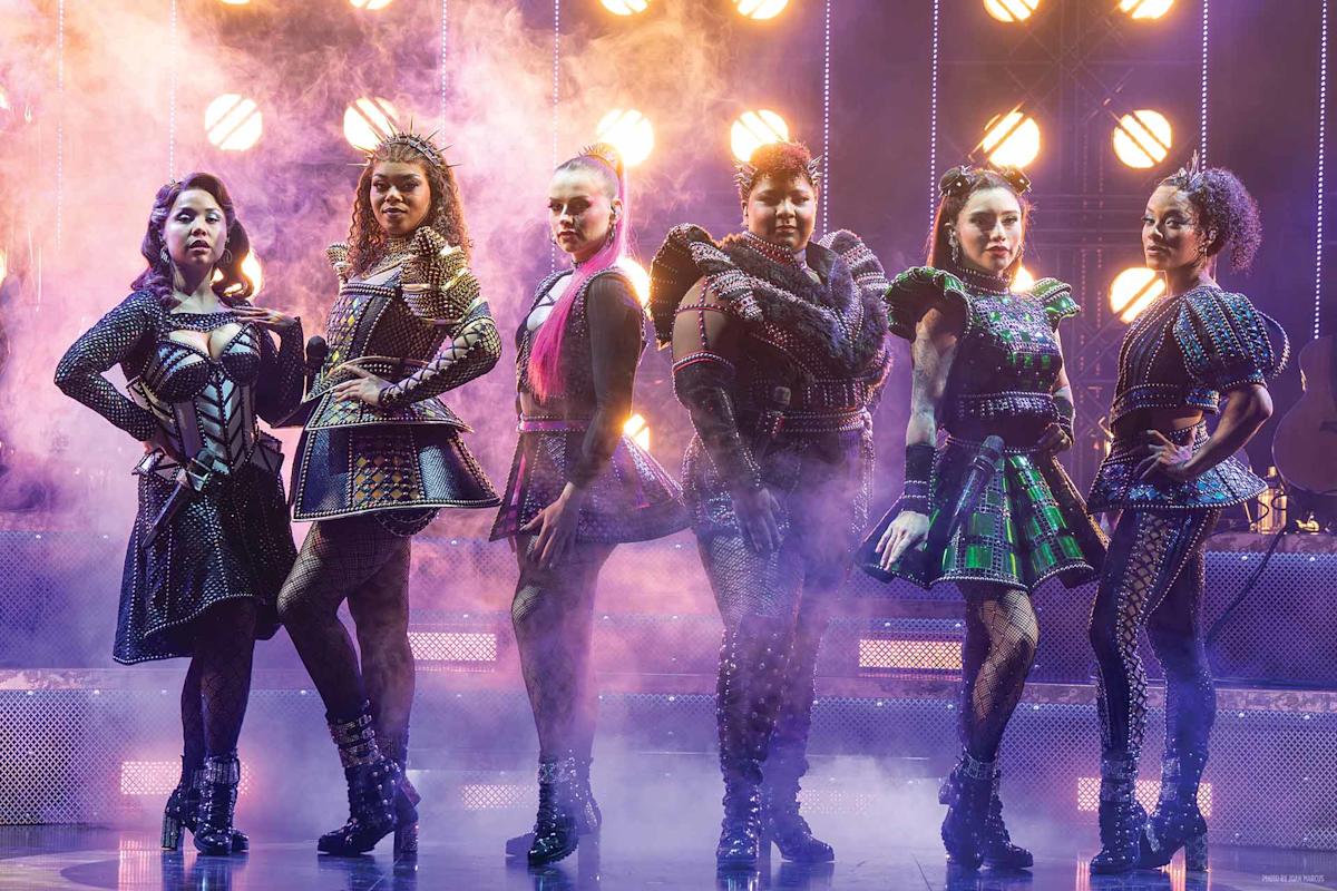 Six women in colorful, ornate costumes pose confidently on a stage with dramatic lighting and smoke effects, evoking a modern, theatrical, and royal atmosphere.