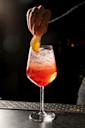 A hand garnishes a bright red cocktail in a wine glass with an orange slice. The drink is topped with ice and the background is dark, highlighting the vibrant colors of the cocktail.