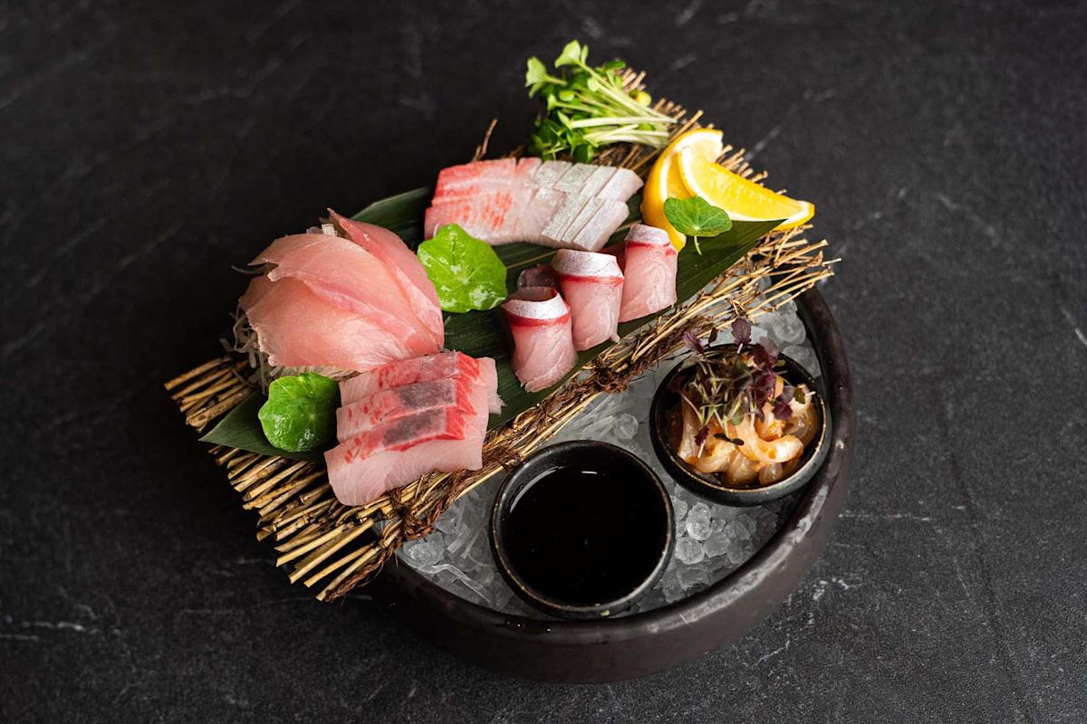 A platter of assorted sashimi with slices of raw fish, lemon wedge, microgreens, wasabi, dipping sauce, and a small side dish, all arranged on a bamboo mat over ice in a black bowl.